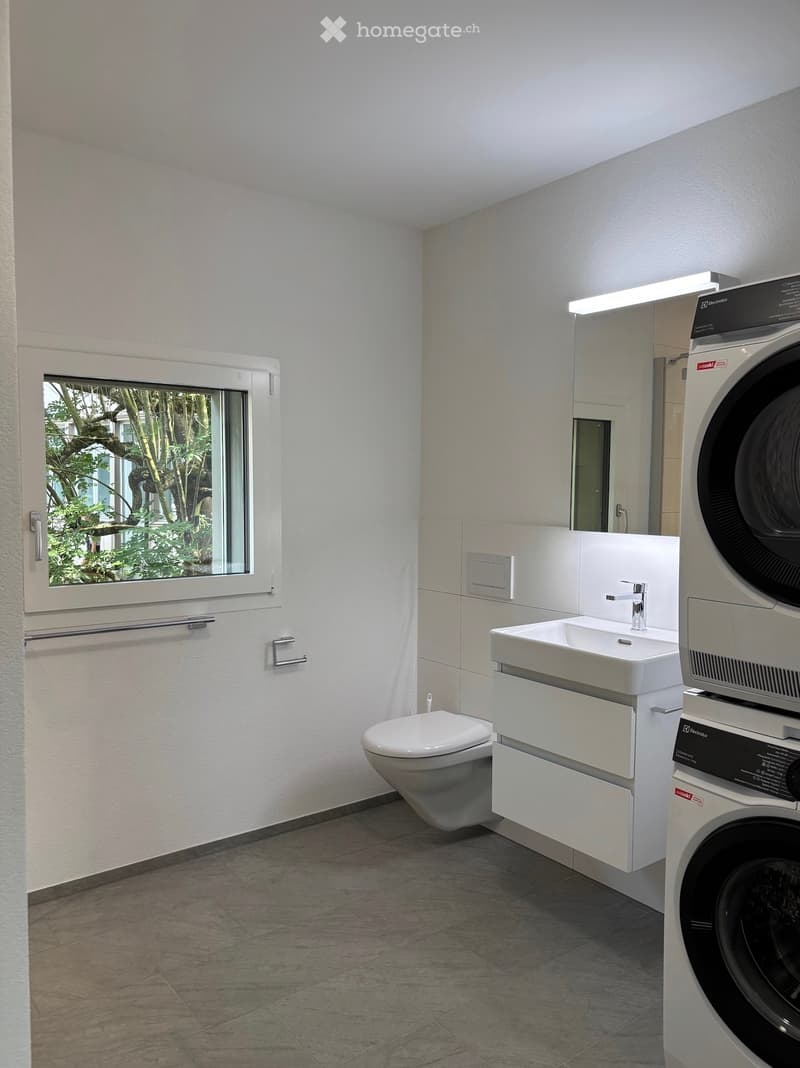 Modern bathroom with a toilet, sink, faucet, washing machine, and a large window offering a view of greenery outside.