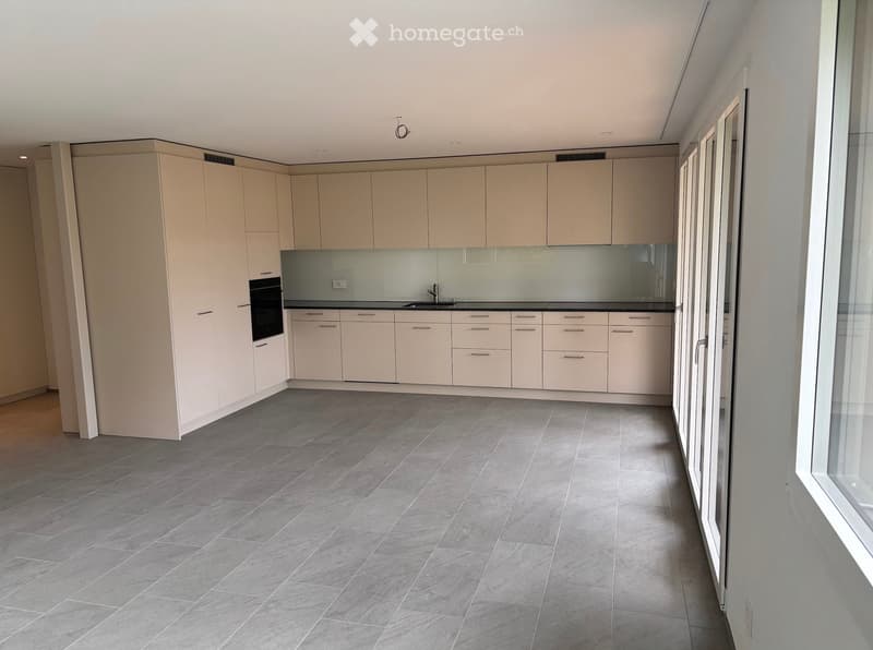 Modern kitchen with light-colored cabinets, a central island, and a dark countertop. The floor is tiled in a gray pattern, and the space is well-lit with recessed lighting.