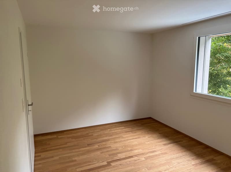 Empty room with wooden flooring, white walls, and a window showing greenery outside.