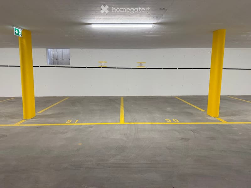 Empty underground parking area with marked parking spaces, yellow pillars, and a green emergency exit sign.