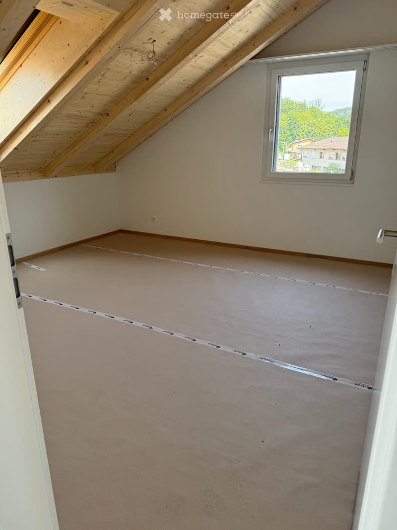 Empty attic room with wooden slanted ceiling, single window, and unfinished walls.