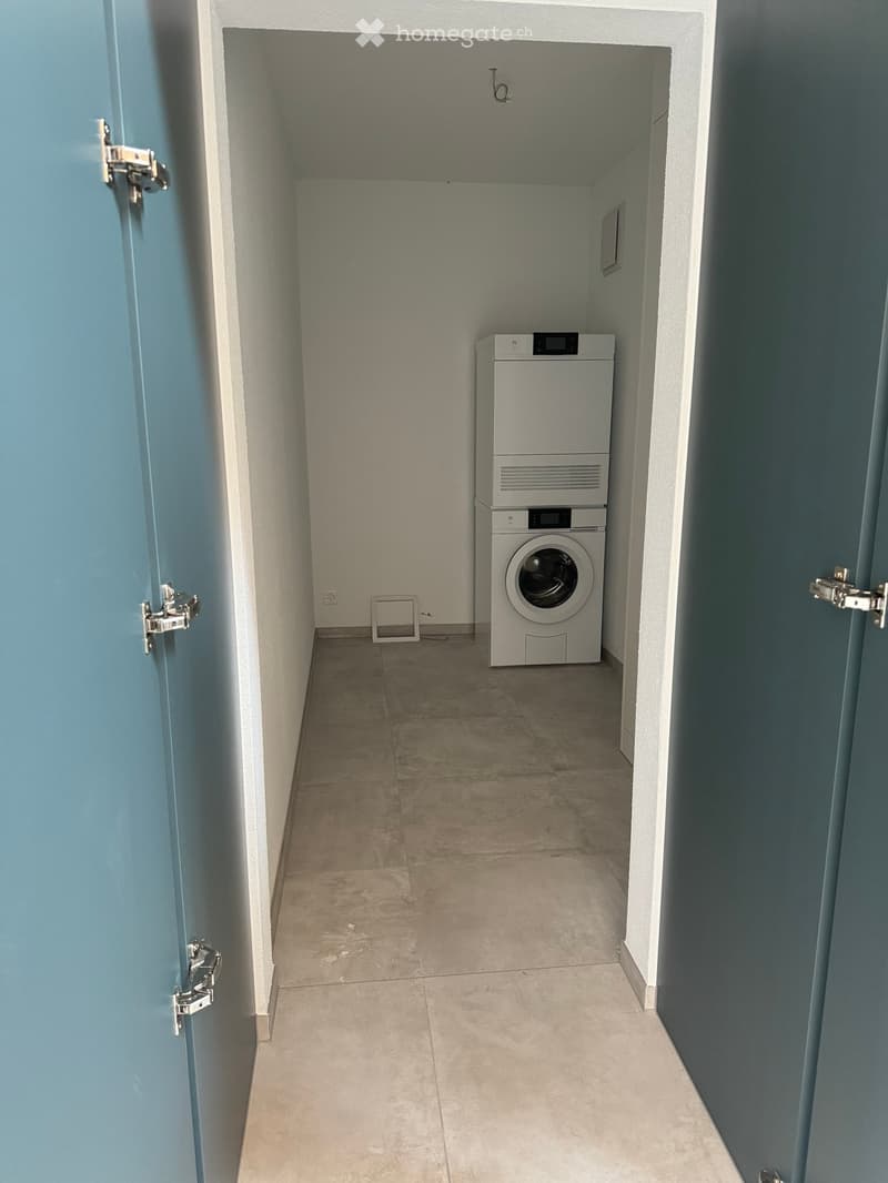 A narrow hallway with tiled flooring and white walls, featuring a washing machine and dryer unit at the end.