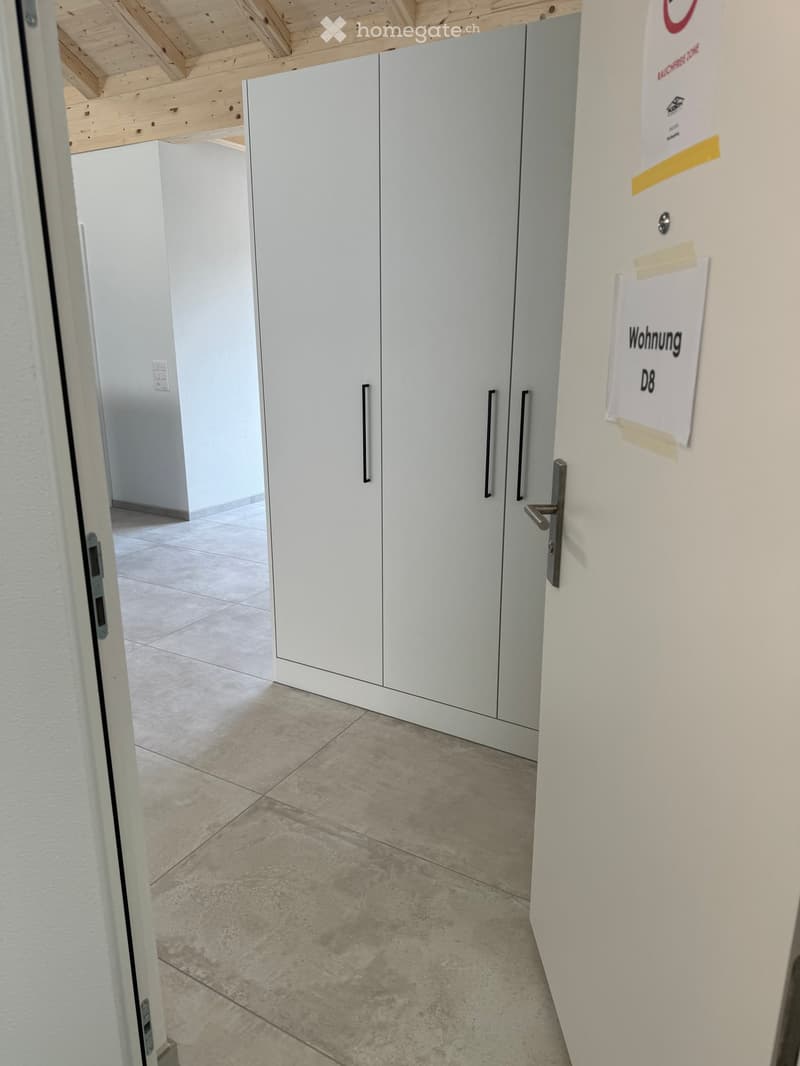 Empty hallway with white walls, tiled floor, and a large white cabinet with black handles. A sign on the wall reads 'Wohnung D8'.