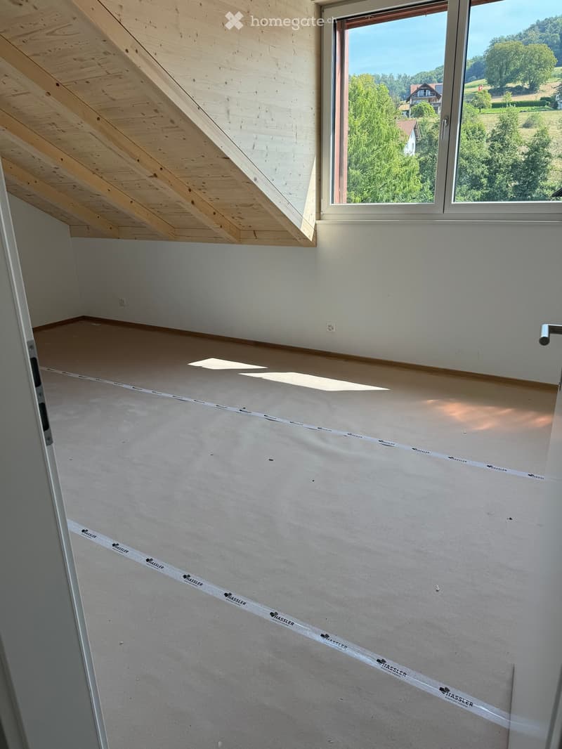 Empty attic room with wooden slanted ceiling, large window showing outdoor view, and protective tape on floor.