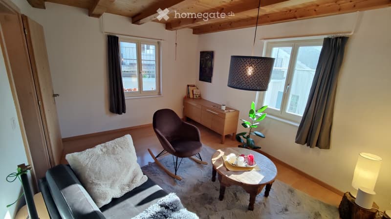 Cozy living room with exposed wooden beams, large windows, a dark grey sofa with a white fur throw, a wooden coffee table, a rocking chair, a sideboard, and a hanging lamp.