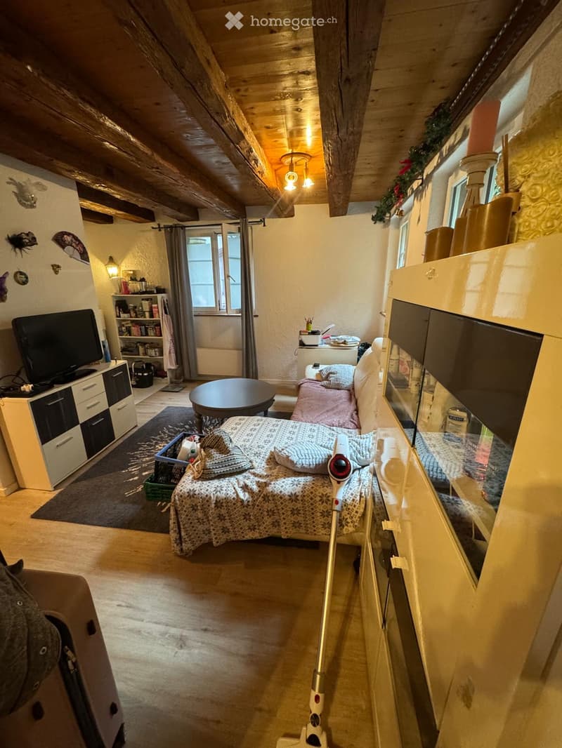 Cozy attic bedroom with exposed wooden beams, a bed with patterned bedding, a small sofa, a round table, a TV cabinet, and shelves with various items.