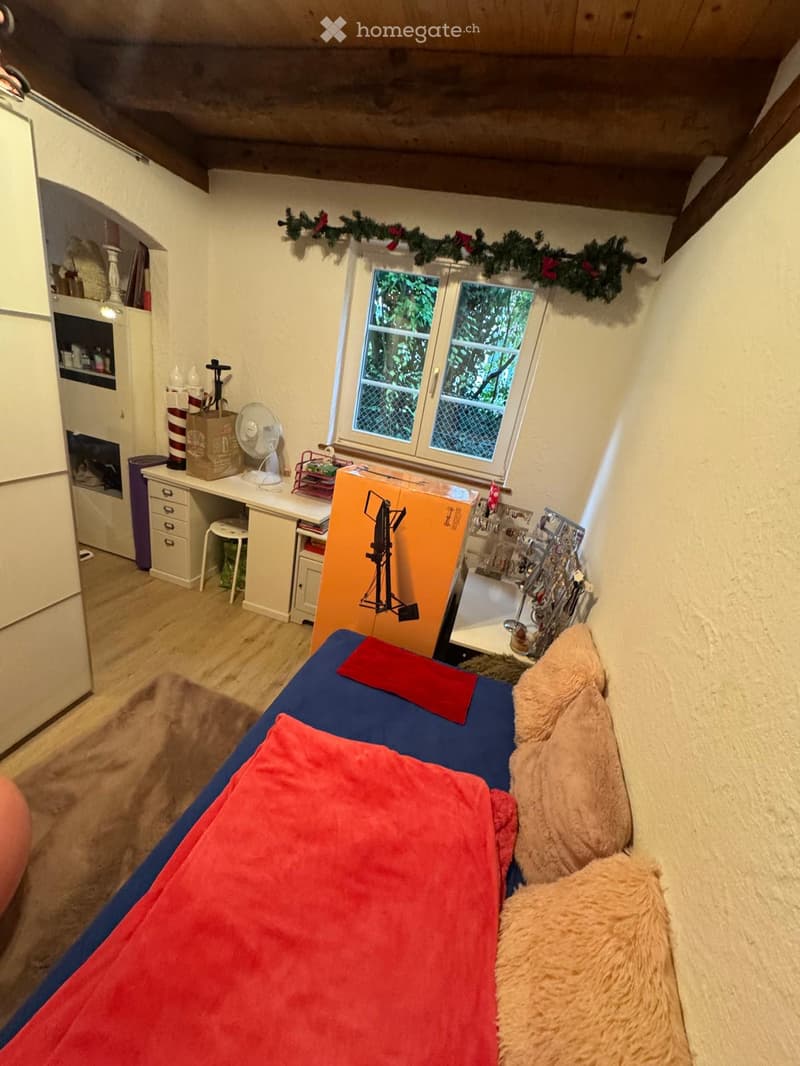 Cozy attic bedroom with slanted wooden ceiling, bed with red and blue bedding, desk with fan and storage, small window with greenery view, and decorative garland above.
