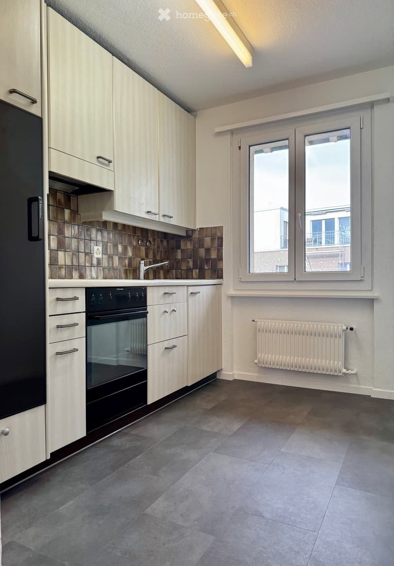 Modern kitchen with light wood cabinets, stainless steel appliances, dark tiled backsplash, and grey tiled flooring.