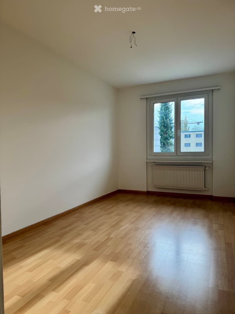 Empty room with wooden flooring, white walls, and a large window with a view of trees and buildings.