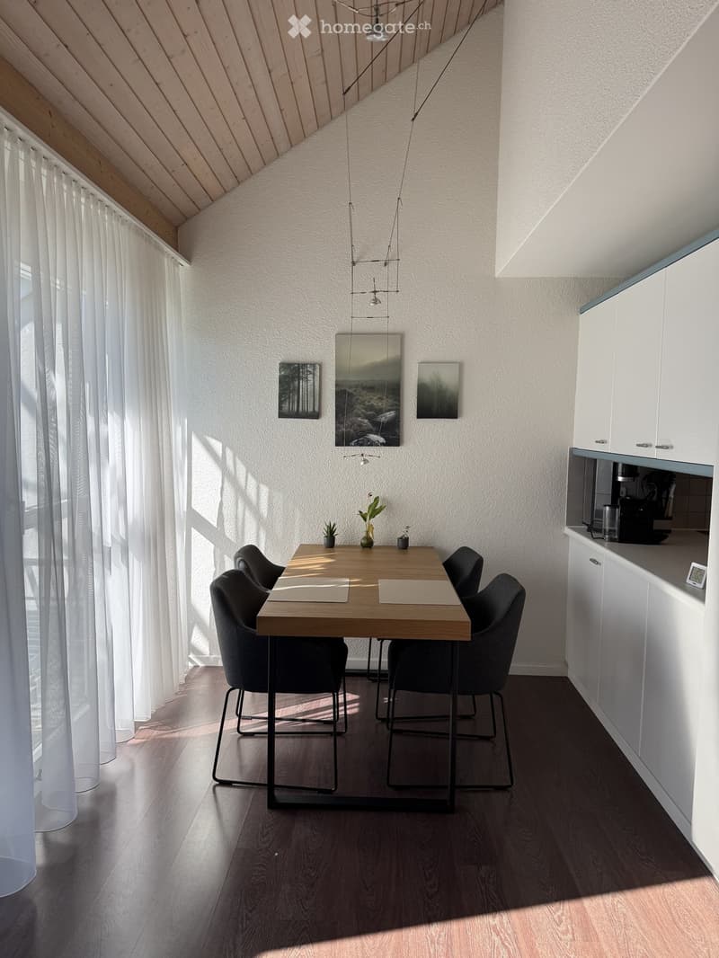 Modern dining area with a wooden table, four black chairs, white cabinets, and a coffee machine on the counter. The room features large windows with white curtains, a pendant light fixture, and several decorative items on the table.