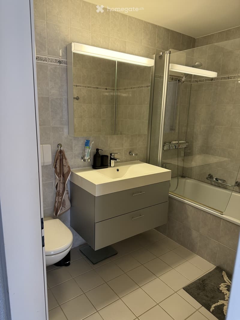 Modern bathroom with a white sink, mirror with built-in lighting, toilet, bathtub, and tiled walls and floor.