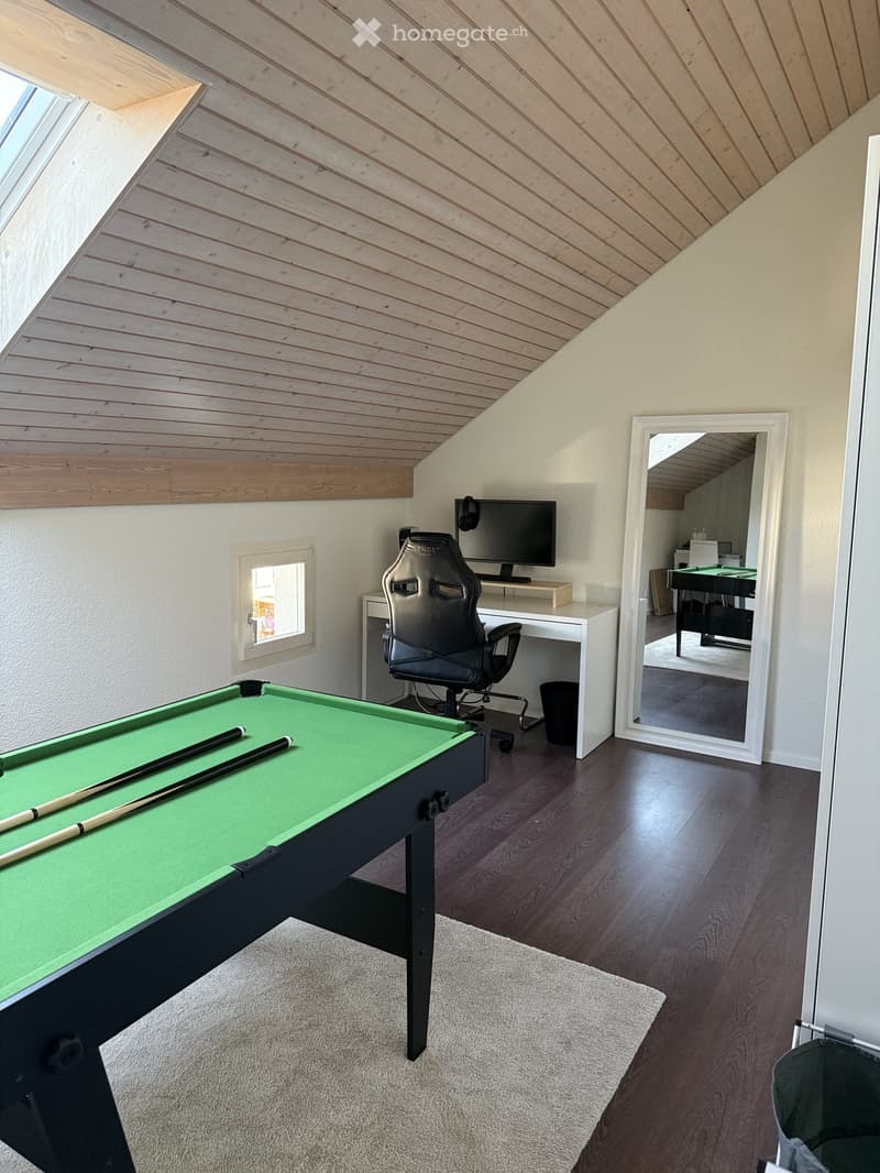 A modern attic room with wooden slatted ceilings, dark hardwood floors, and a pool table in the foreground. A desk with a computer monitor and a gaming chair is positioned near a window, and a large mirror reflects part of the room.