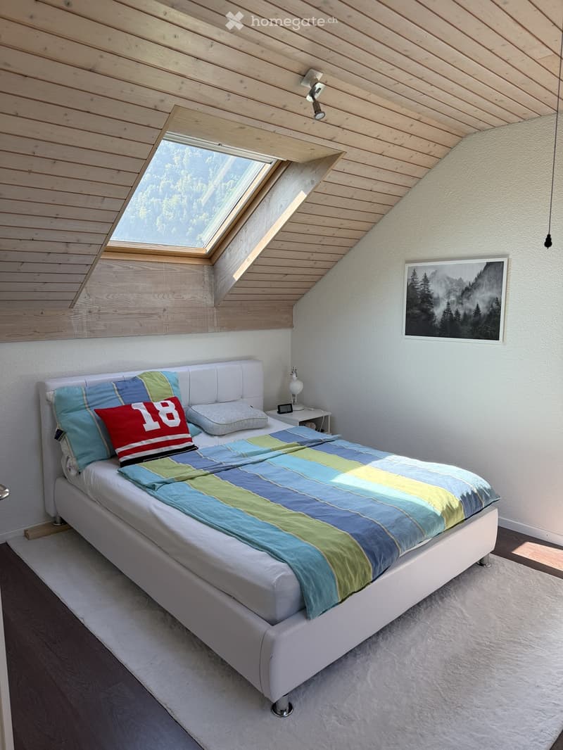 Modern bedroom with a white bed, colorful bedding, a nightstand with a lamp, and a picture frame on the wall. The room features wooden ceilings and a skylight window.