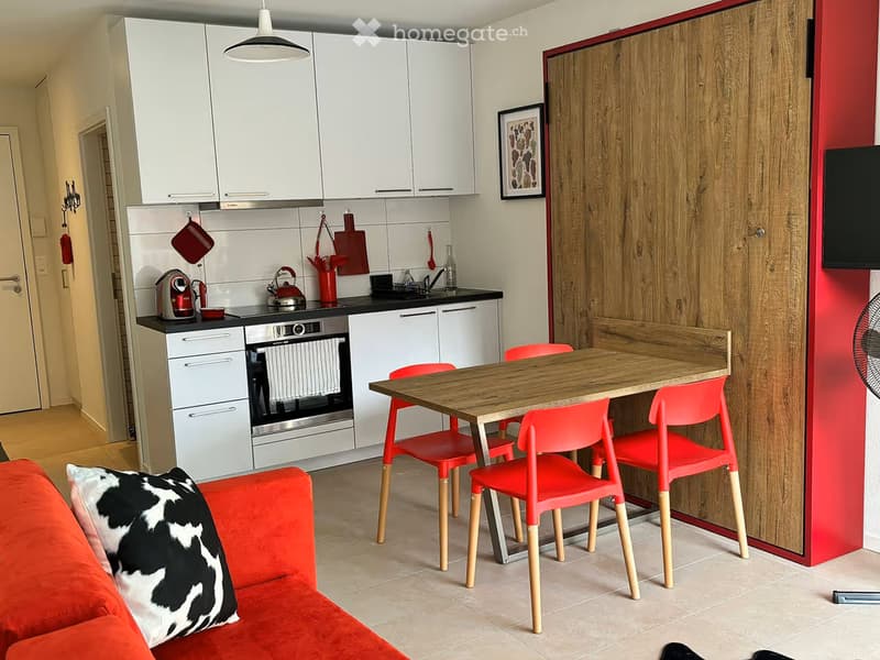 Modern open-plan living and dining area with a red sofa, a wooden dining table with red chairs, a kitchen area with white cabinets, an oven, and a fridge.