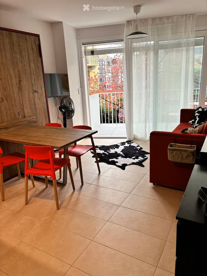 A modern living room with a red sofa, a wooden dining table with red chairs, a cowhide rug, and a fan near the TV screen. The room has large windows with sheer curtains and a view of a balcony.