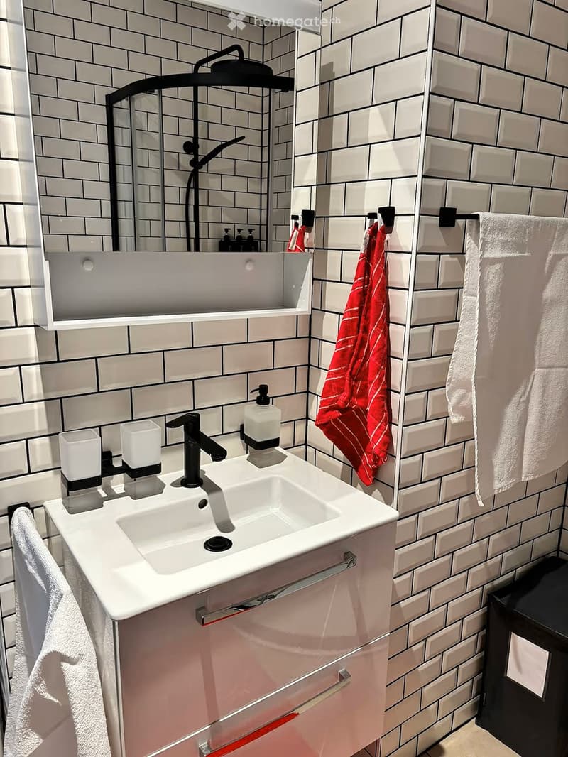 Modern bathroom with white tiled walls, a white sink with a black faucet, a shower head, and two towels hanging on a rack.