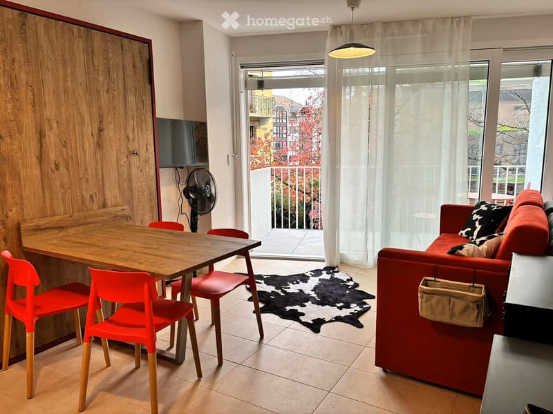A modern living room with a red sofa, wooden dining table, red chairs, a fan, and a view of a balcony with outdoor furniture.