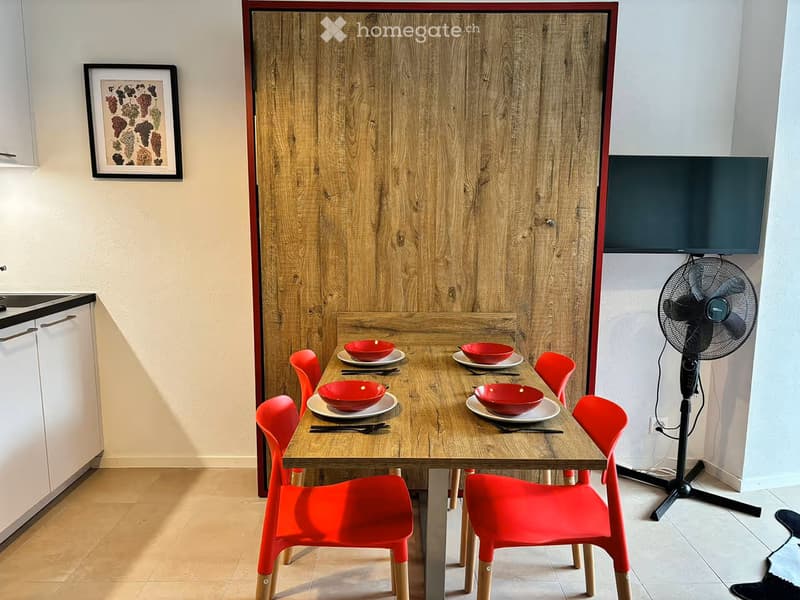 A modern dining area with a wooden table, red chairs, and a fan. The table is set with red bowls and plates, and there is a screen mounted on the wall.