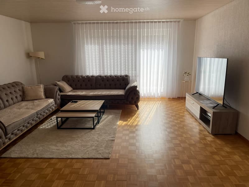 Modern living room with wooden flooring, large windows with white curtains, a grey sectional sofa, a coffee table, a television on a stand, and a floor lamp.