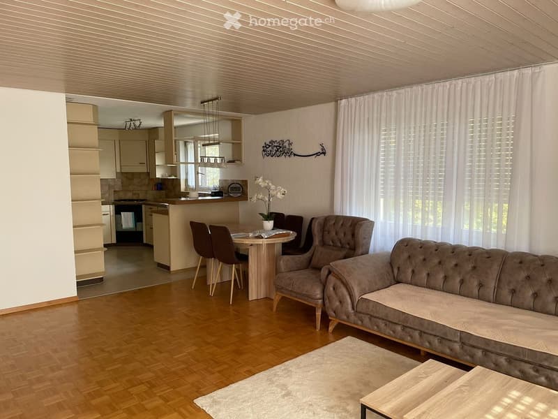 Modern living room with a grey sofa, armchair, coffee table, dining table, and chairs. The room features a kitchen area with cabinets and an oven, and is decorated with a flower vase and artwork on the wall.
