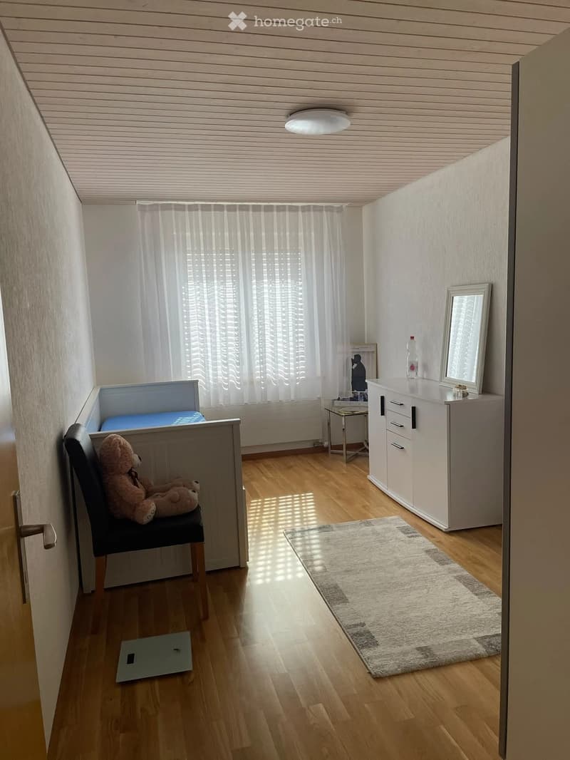A small, well-lit room with wooden flooring, a bed with a blue mattress, a sofa with a teddy bear, a white cabinet, a mirror, a small table, and a rug.