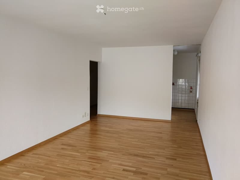 Empty room with wooden flooring and white walls, featuring a doorway and a glimpse of a tiled bathroom area.