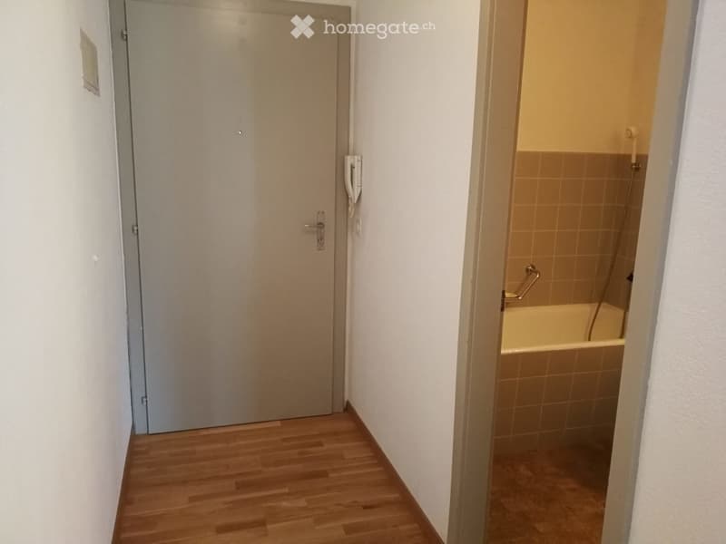 A narrow hallway with wooden flooring, a closed door on the left, and an open doorway leading to a small bathroom with a bathtub and tiled walls.