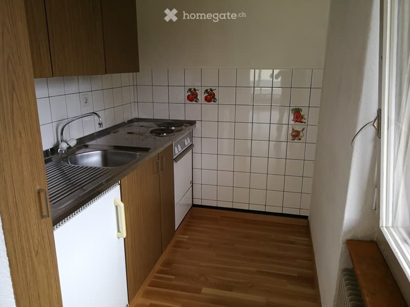 Compact kitchen with wooden cabinets, white tiled backsplash featuring fruit and vegetable designs, a sink with a faucet, a stove, and an oven. The floor is made of light wooden planks.