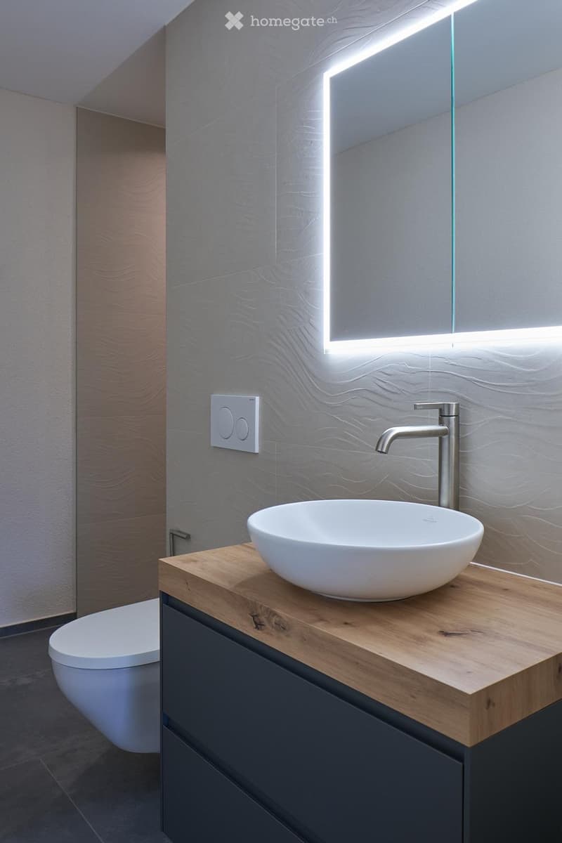 modern bathroom with gray tiled floor, large mirror, oval white sink, wooden countertop, chrome faucet, white toilet, illuminated mirror, tiled walls