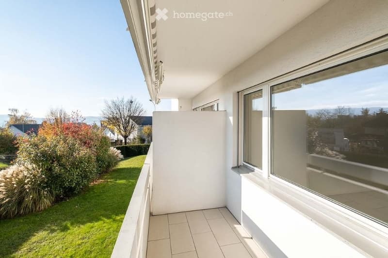 Balcony with tiled floor, large windows, and a view of a green garden and distant landscape.