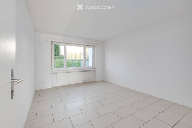 Empty room with white walls, tiled floor, and large window with a view of greenery outside.