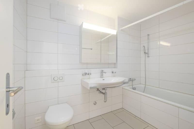 Modern bathroom with white tiles, a sink, toilet, bathtub, and a mirror with built-in lighting.