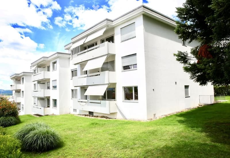 Modern white apartment building with multiple balconies and large windows, surrounded by a well-maintained lawn and trees.