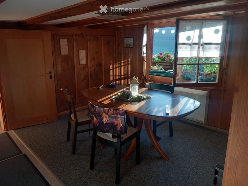 Cozy wooden-paneled room with a round wooden table, chairs, and a decorative lantern on the table. Large windows with white curtains let in natural light, and the room features exposed wooden beams on the ceiling.