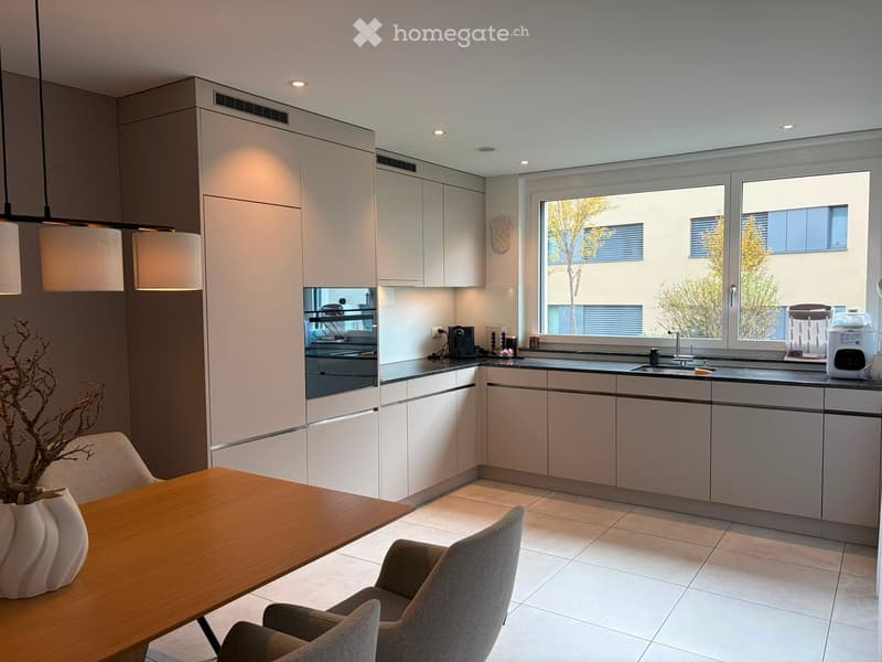 Modern kitchen with white cabinets, a wooden dining table, and grey chairs. Features include a sink, coffee maker, oven, and a blender on the counter.