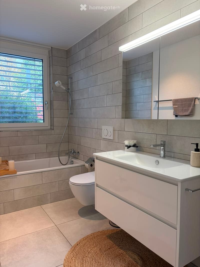 Modern bathroom with a bathtub, toilet, sink, and large mirror. Features tiled walls, a window with blinds, and a wicker basket on the floor.