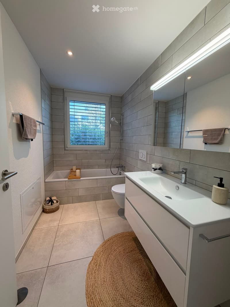 Modern bathroom with a large sink, toilet, bathtub, and a window with blinds. Features include a wicker rug, a wicker basket, and a candle on the bathtub ledge.