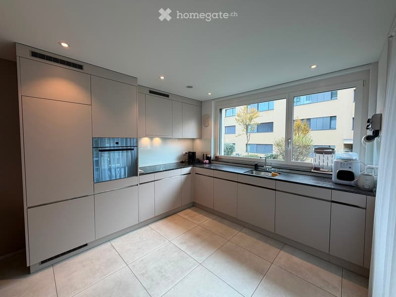 Modern kitchen with light grey cabinets, a built-in oven, a sink with a faucet, and various small appliances on the counter, including a coffee maker, toaster, and blender. The space is well-lit with recessed lighting and has large windows offering a view