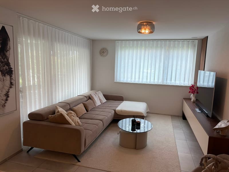 Modern living room with a beige sectional sofa, glass coffee table, TV stand, and decorative vase on the stand. Large windows with vertical blinds and a wall-mounted clock are visible.