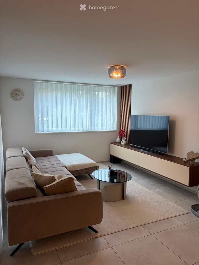 Modern living room with a beige sectional sofa, glass coffee table, flat-screen TV on a wooden cabinet, and decorative vases. The room features large windows with blinds and a stylish chandelier.