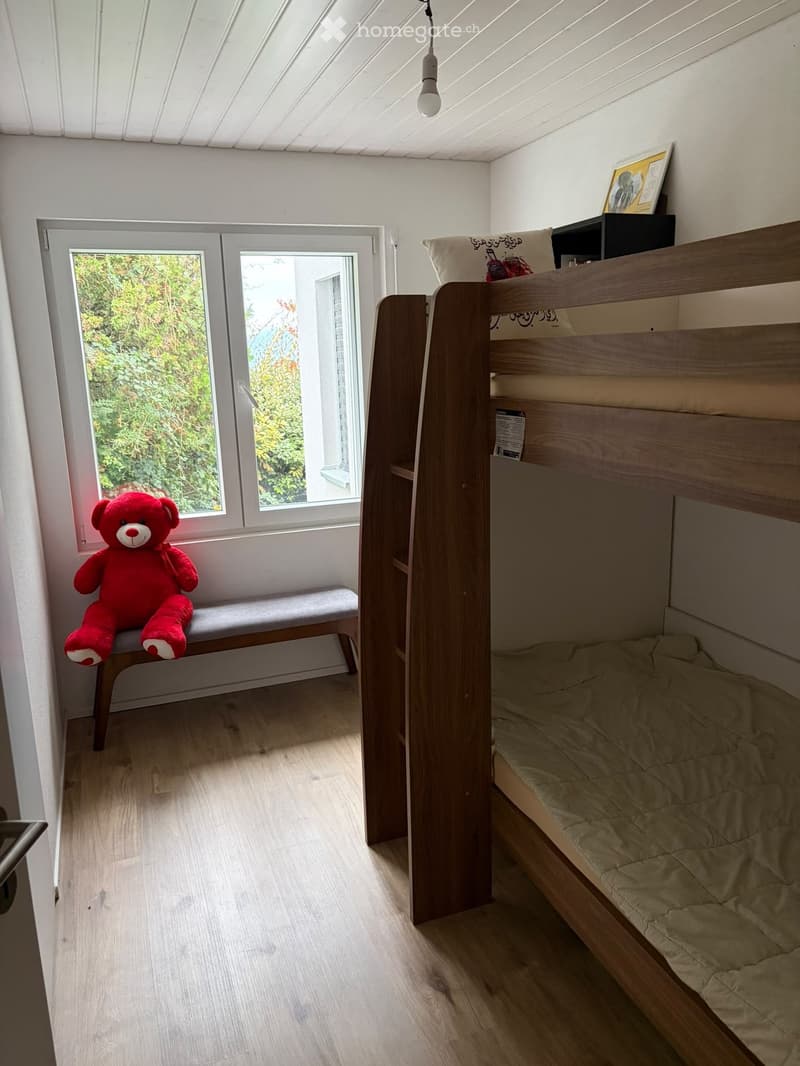 Cozy bedroom with a wooden bunk bed, a small bench with a red teddy bear, and a window with a view of greenery.