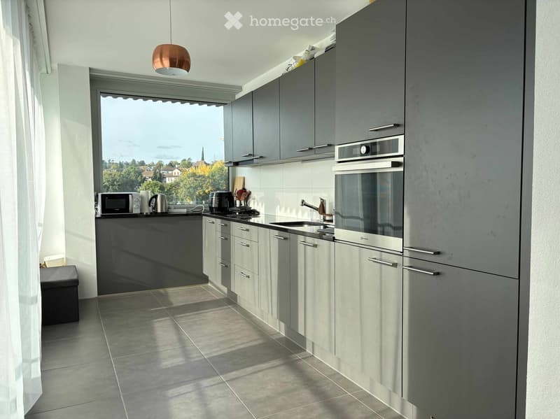 Modern kitchen with grey cabinets, stainless steel appliances, and a large window offering a scenic view. Includes a microwave, oven, sink, and various kitchen items on the counter.