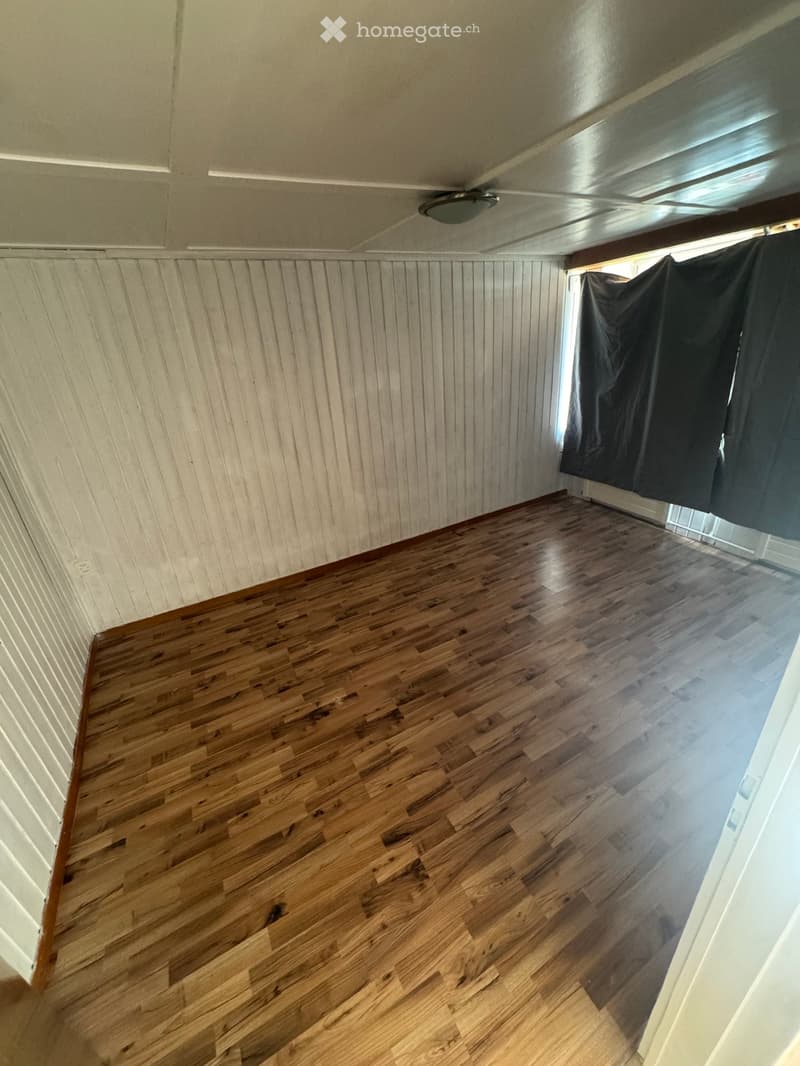 Empty room with wooden flooring and white paneled walls, featuring a ceiling light fixture and a window with black curtains.