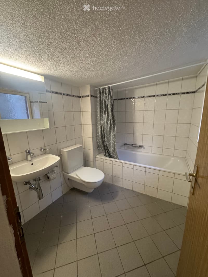 Modern bathroom with white tiles, a sink, toilet, bathtub, and a shower curtain. Features a mirror above the sink and a window with frosted glass.