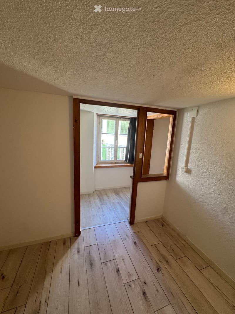 Empty hallway with wooden flooring, white walls, and a doorway leading to another room with a window and balcony view.