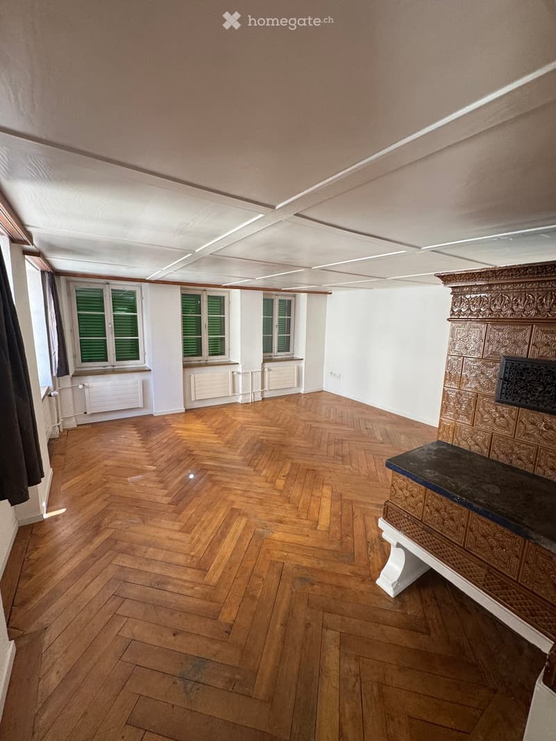 Empty room with wooden herringbone flooring, white walls, and a decorative fireplace with ornate carvings. Windows with green shutters and radiators are visible.