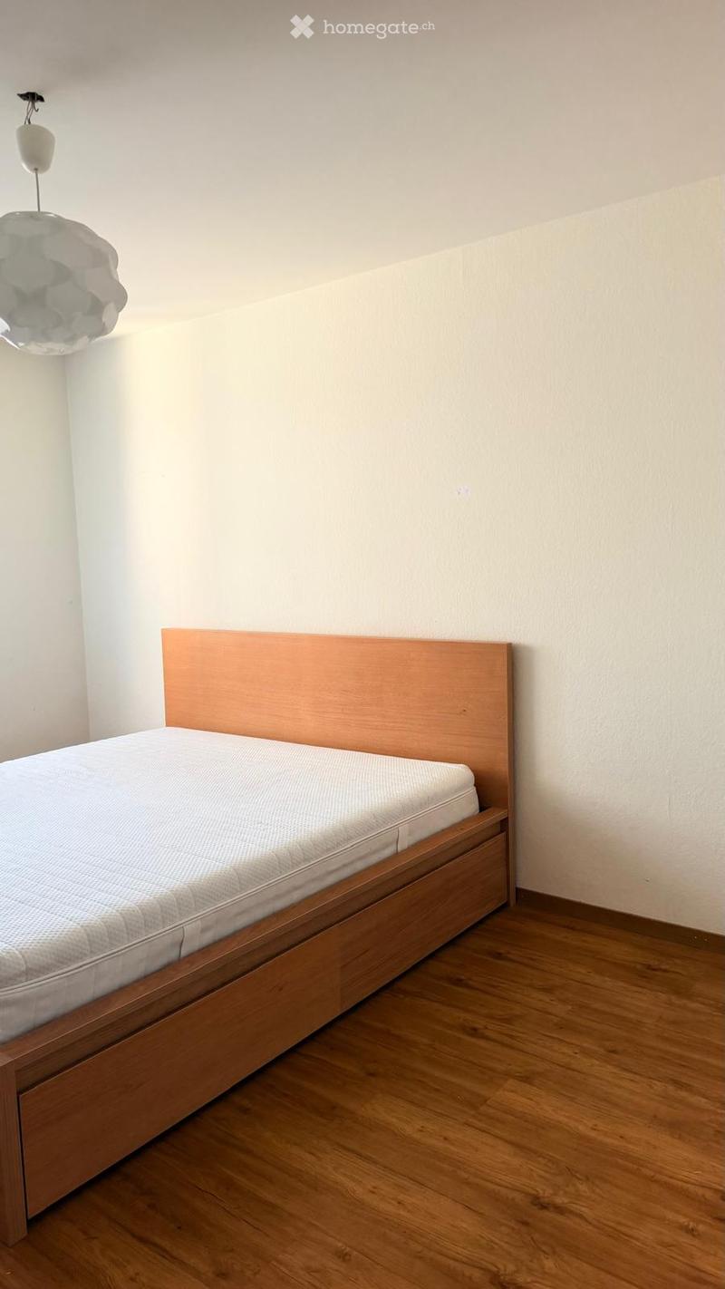 A simple bedroom with a wooden bed frame, a mattress, and a headboard. The room features light-colored walls and a wooden floor.