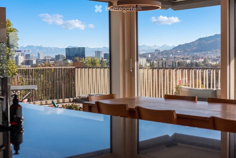 Modern dining area with a wooden dining table and chairs, a sofa on the balcony, and a panoramic city view with mountains in the background.
