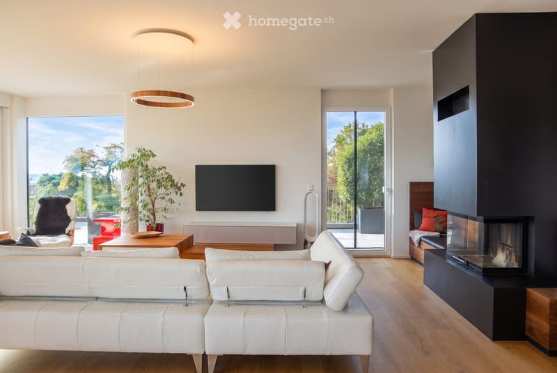 Modern living room with white leather sofa, coffee table, fireplace, TV, large windows, and a view of greenery outside.