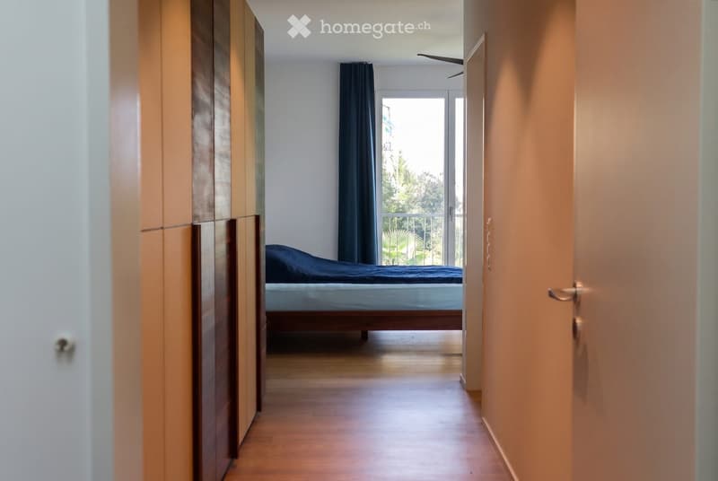 A modern bedroom with a wooden floor, a bed with blue bedding, a wardrobe, and a large window with dark curtains leading to a balcony.